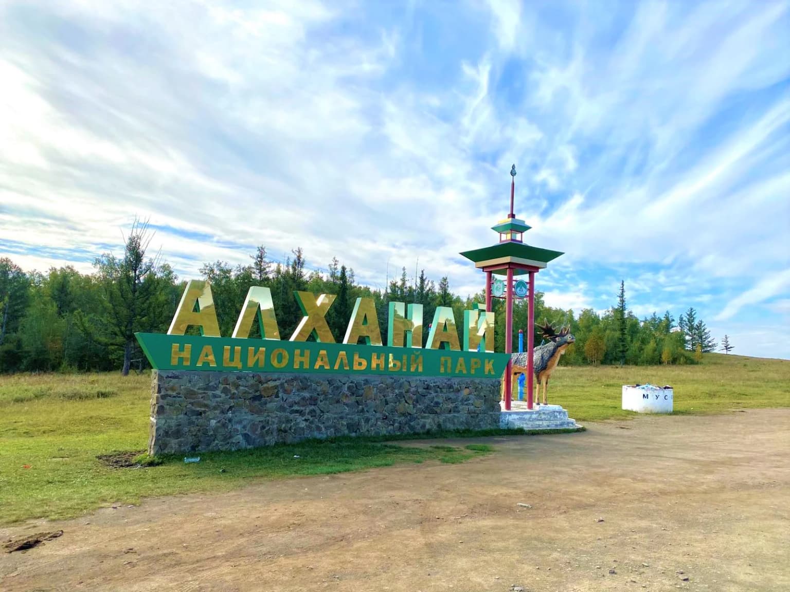 Stone and metal sign reading 'АЛХАНАЙ НАЦИОНАЛЬНЫЙ ПАРК' (Alkhana National Park) in Russian, with a traditional structure and forest background