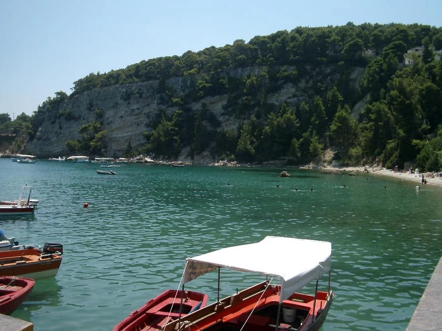 Several boats docked at a waterfront with a rocky cliffside and forested hillside in the background