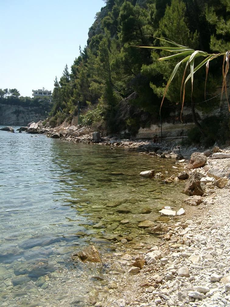 Clear blue water along a rocky shore with forested hills in the background under a sunny sky