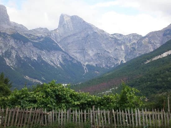 Mountain landscape featuring Maja e Arapit peak, green valley, wooden fence, and forested slopes under a partly cloudy sky