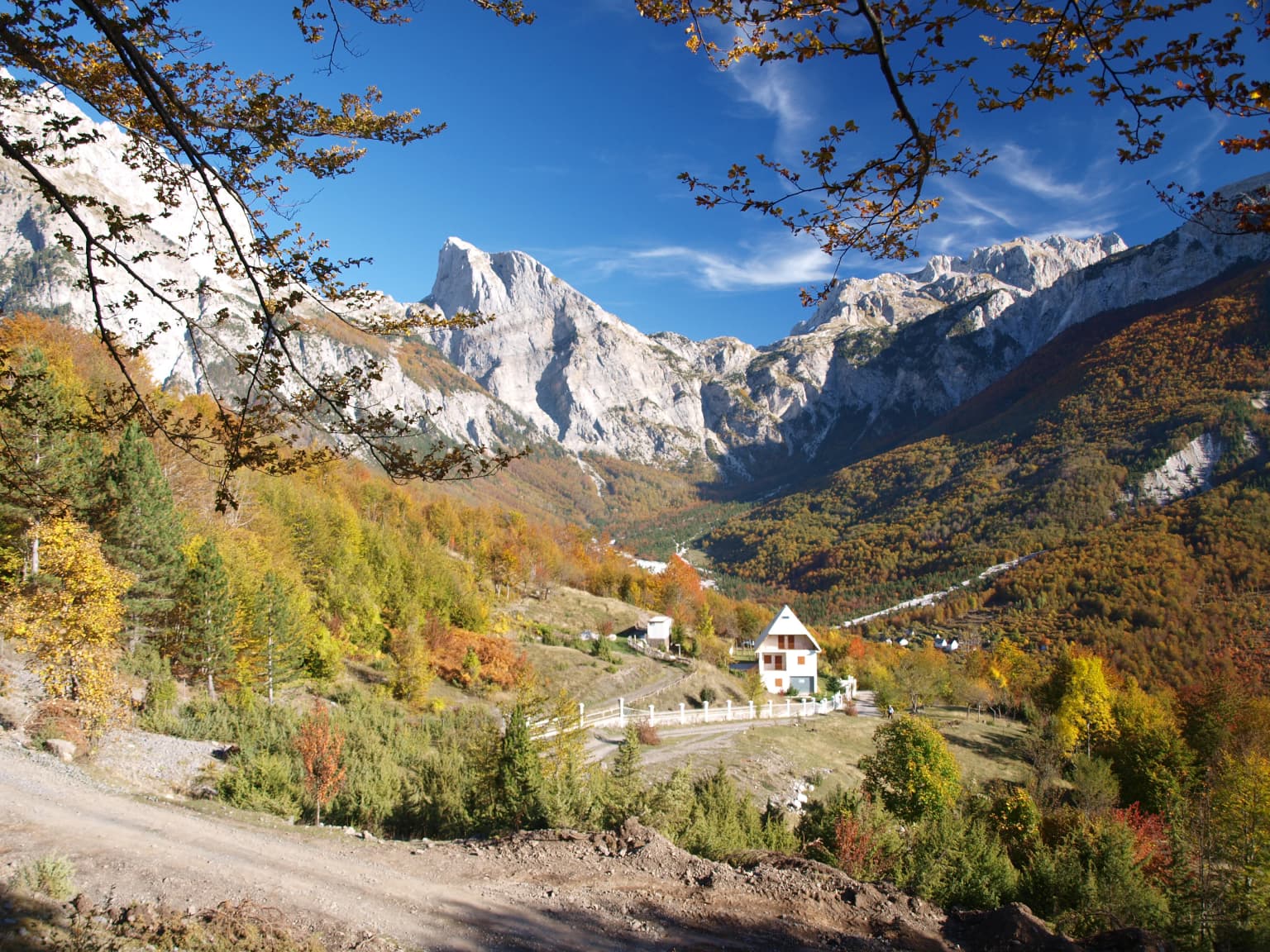 Mountain landscape with autumn foliage, a village with a white house, and rocky peaks under a clear blue sky