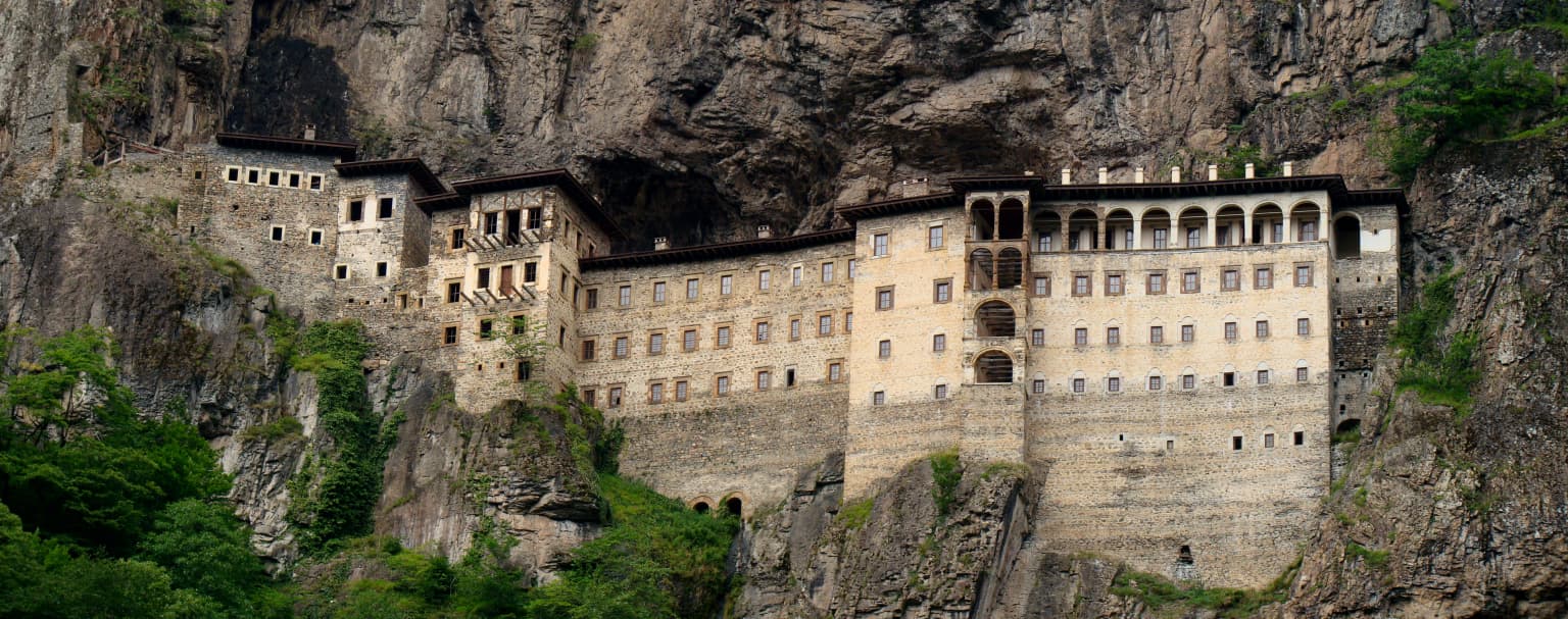 Stone monastery built into a cliffside with lush green vegetation, viewed from a valley across rocky terrain