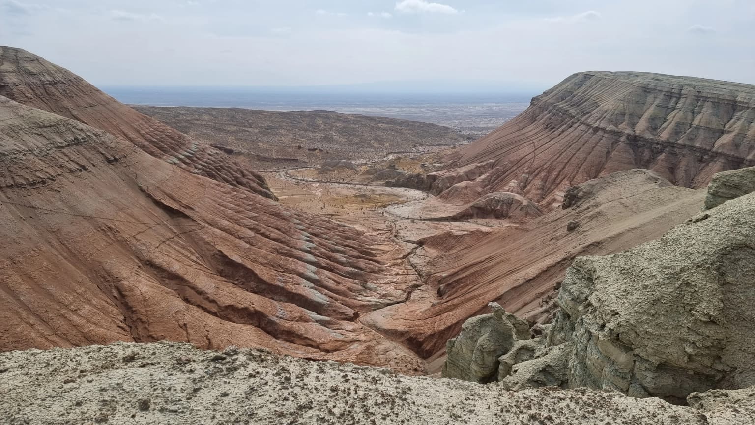 Panoramic view of a deep gorge with red and brown rock layers, dry riverbed, and rocky terrain under a partly cloudy sky