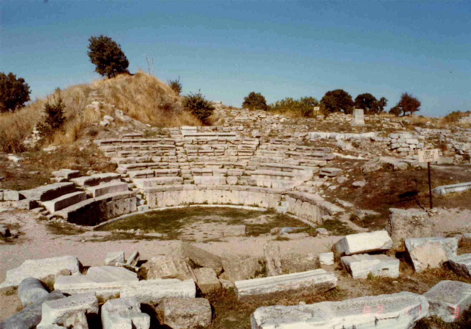 Stone amphitheater ruins with tiered seating, grassy center, and scattered stone blocks under clear sky