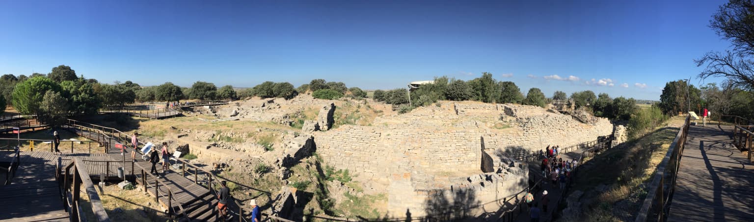 Panoramic view of ancient stone walls from Troy's Level VI site, with visitors on pathways, trees, and clear blue sky