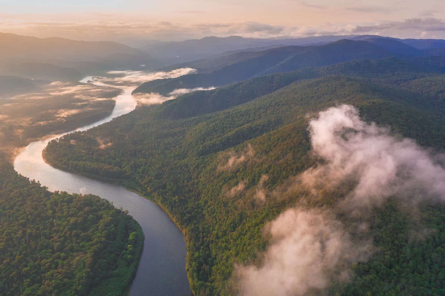Aerial view of a winding river surrounded by dense green forest and mountains with low-lying clouds