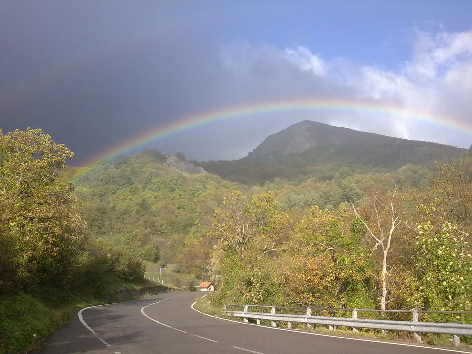 Winding road with guardrail, surrounded by trees and hills, with a rainbow arching over a mountain peak under partly cloudy sky