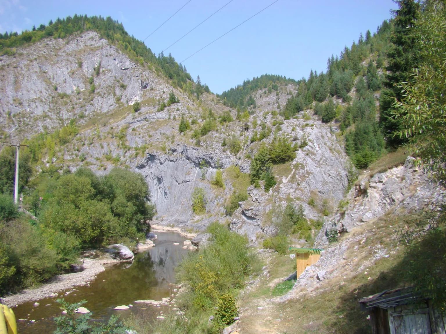 River flowing through a mountain valley with rocky cliffs, forested slopes, and a small structure on the right side