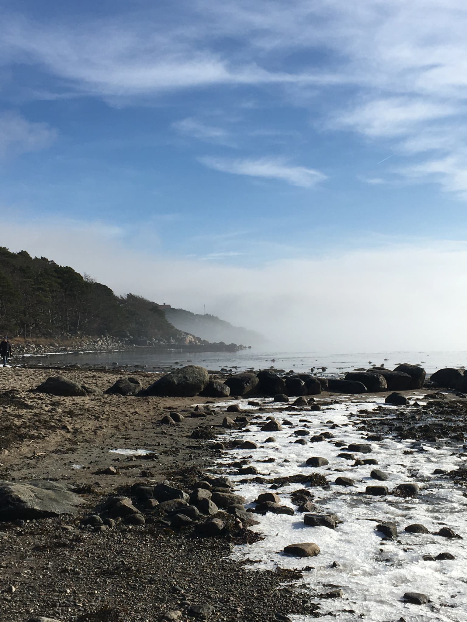 Rocky beach with patches of snow, fog-covered sea, forested hillside, and a person walking under partly cloudy sky