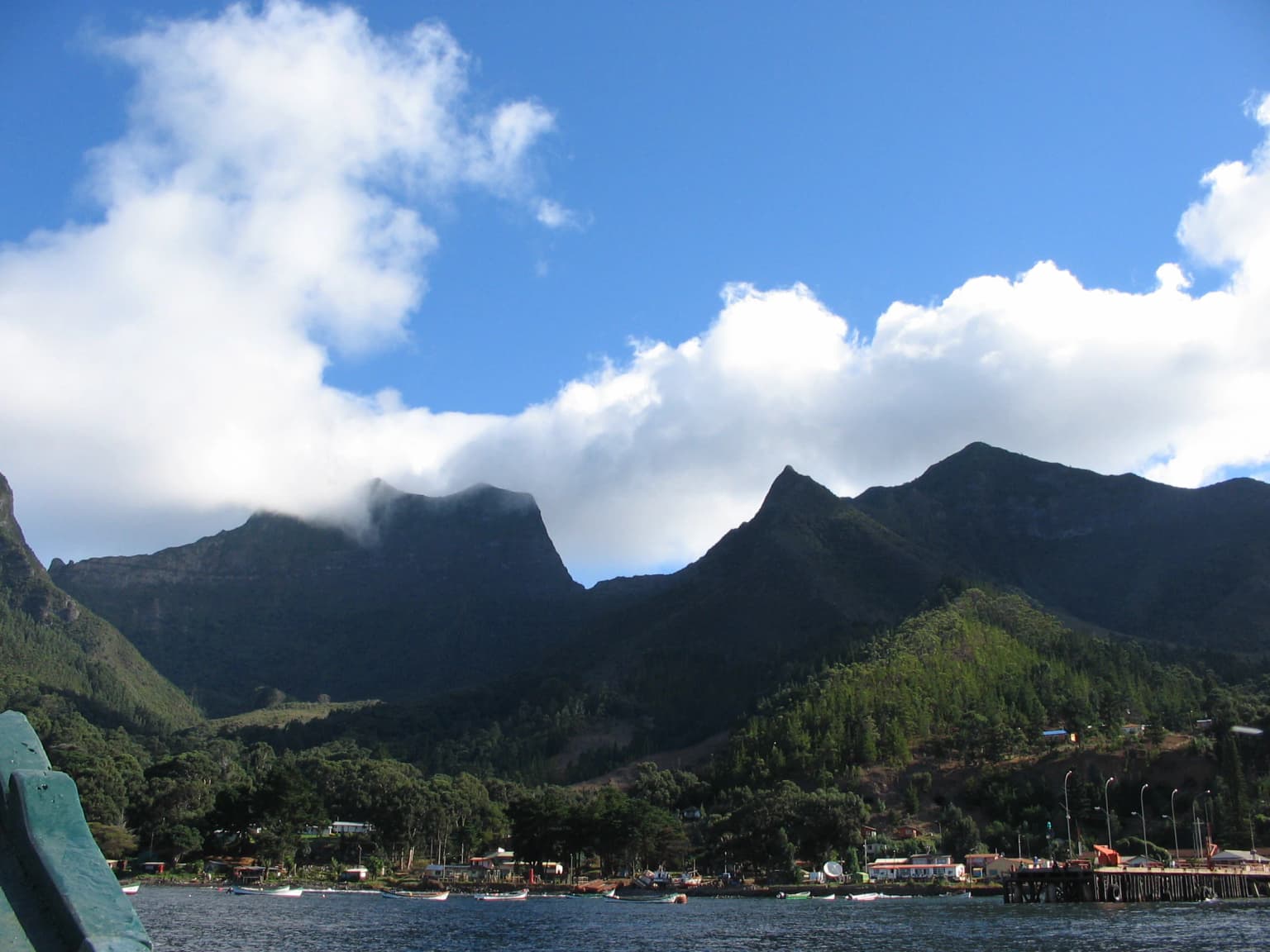 Mountainous landscape of Robinson Crusoe Island with green hills, a body of water, and coastal buildings under a partly cloudy sky