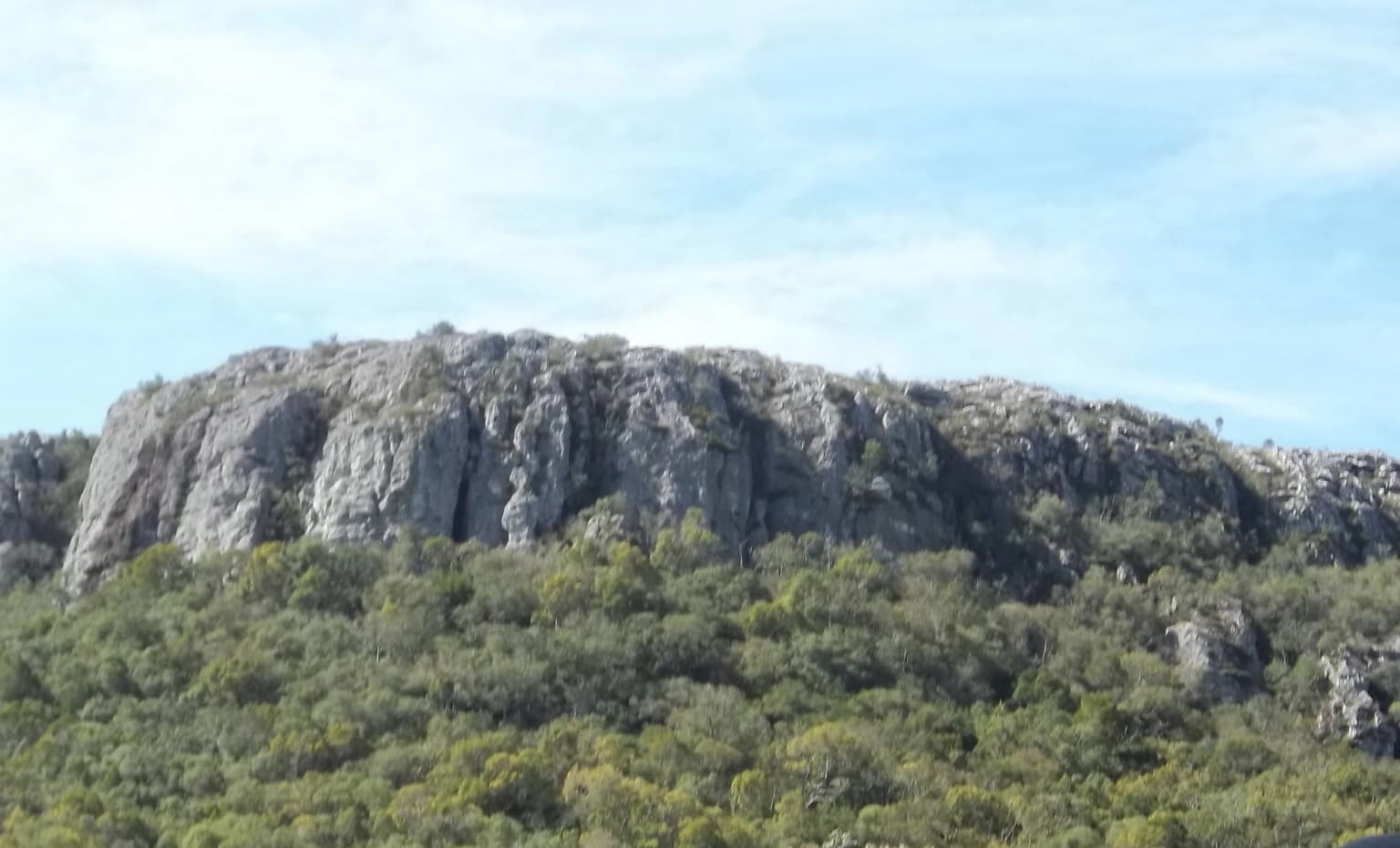 Una gran colina rocosa con una base boscosa bajo un cielo parcialmente nublado