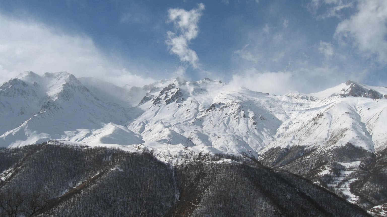 Snow-covered mountain peaks under a partly cloudy sky with forested slopes in the foreground