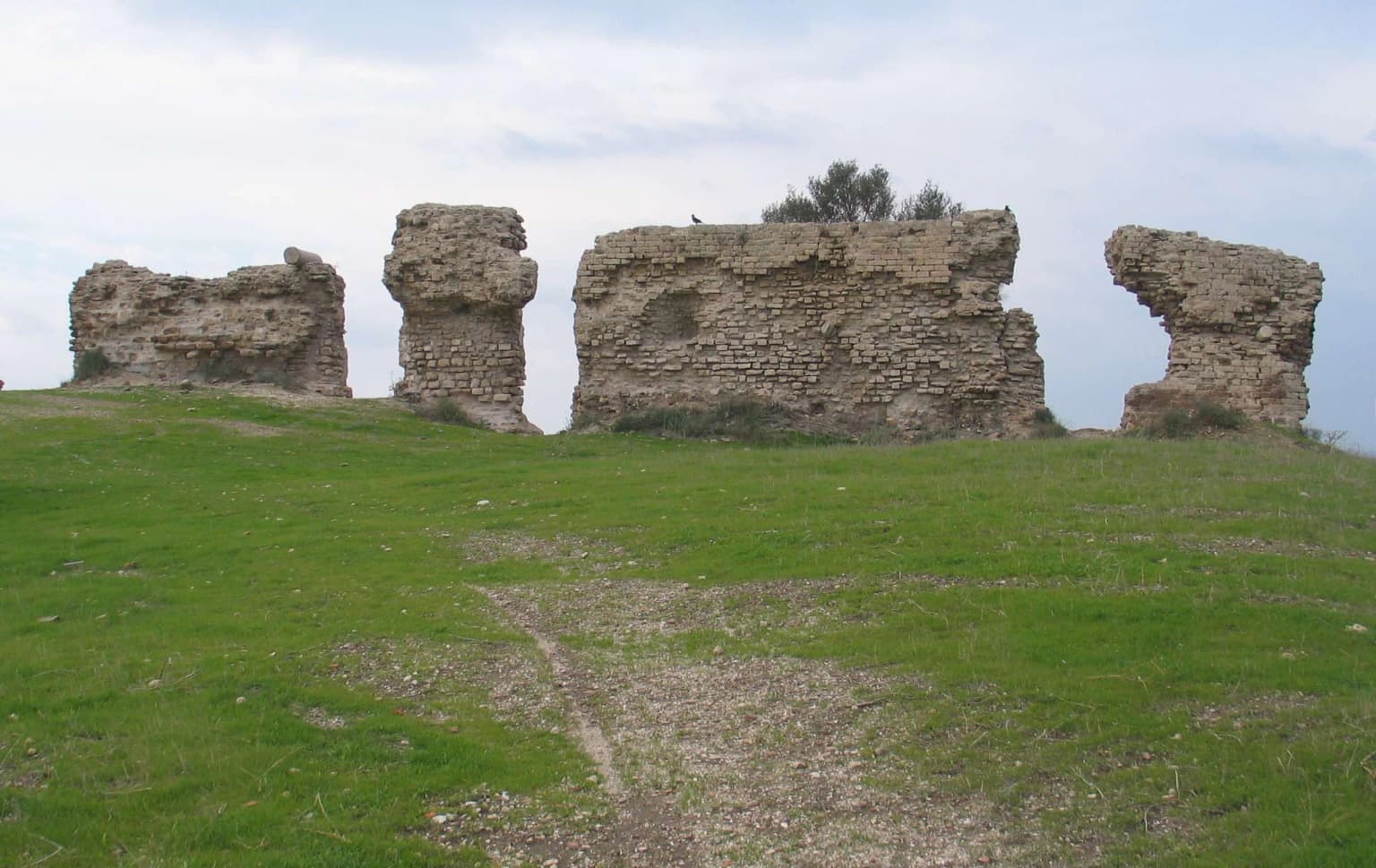 Ruinas de piedra en una colina cubierta de hierba con un sendero de tierra que conduce a ellas bajo un cielo parcialmente nublado.