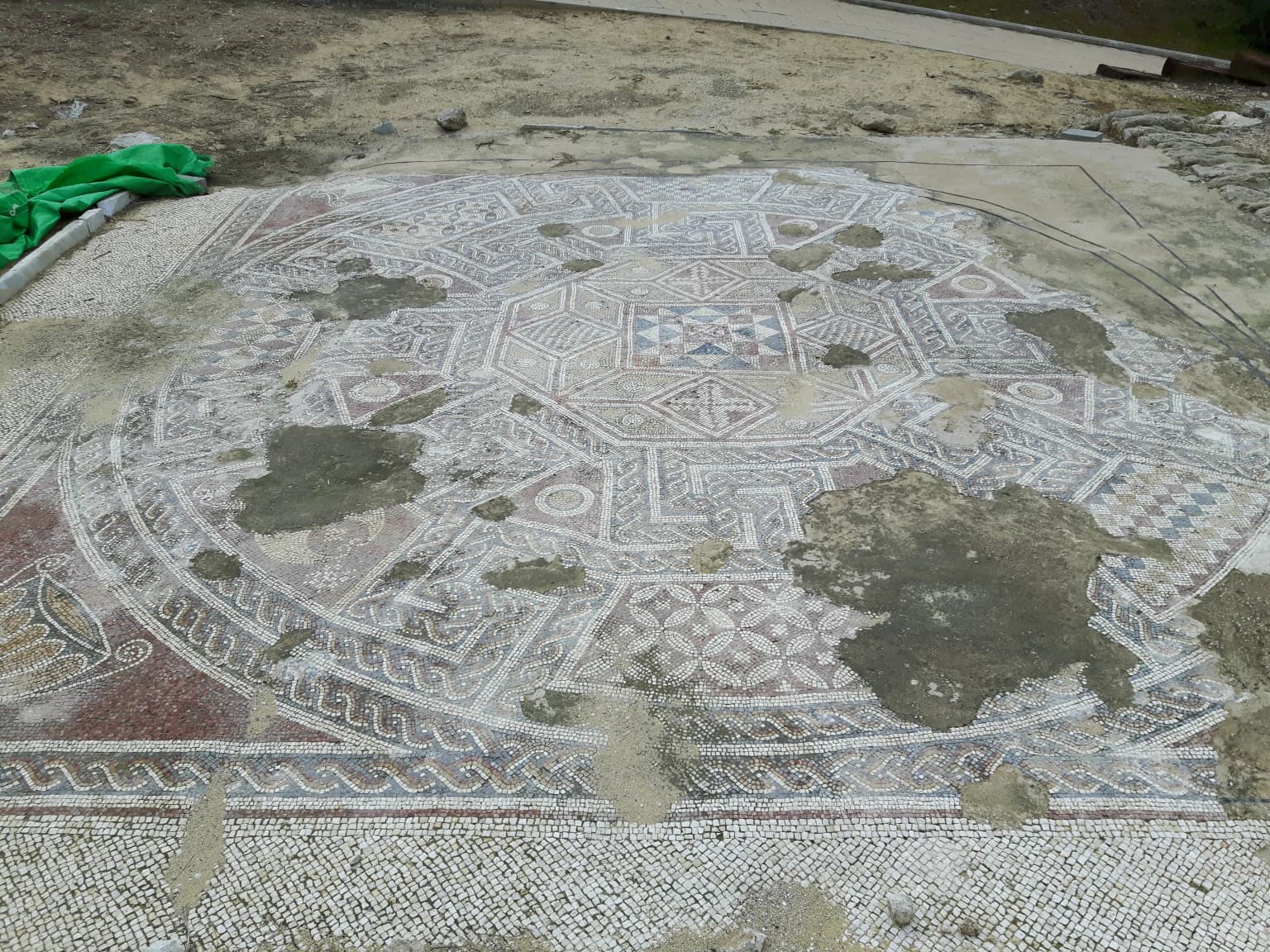 Un gran suelo de mosaico circular con motivos geométricos y zonas de baldosas faltantes, situado en un yacimiento arqueológico