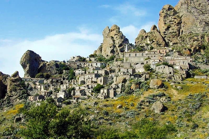 Stone village buildings on a rocky hillside with large rock formations and greenery under a clear blue sky