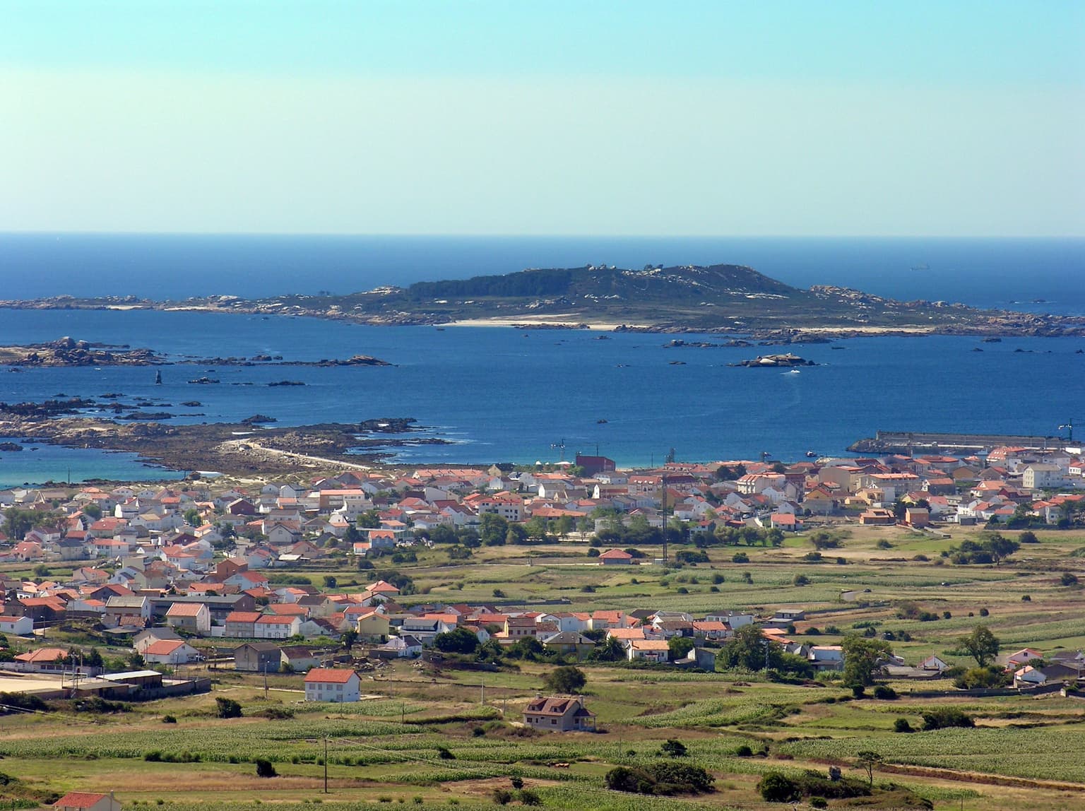 Coastal landscape showing Sálvora Island in the distance, with Aguiño village and surrounding fields in the foreground, overlooking the sea under a clear sky
