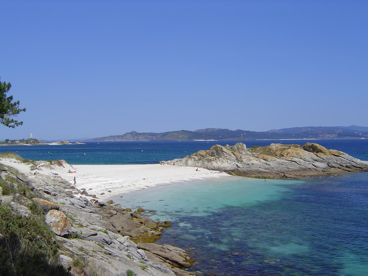 White sand beach with turquoise water, rocky coastline, and distant islands under clear blue sky