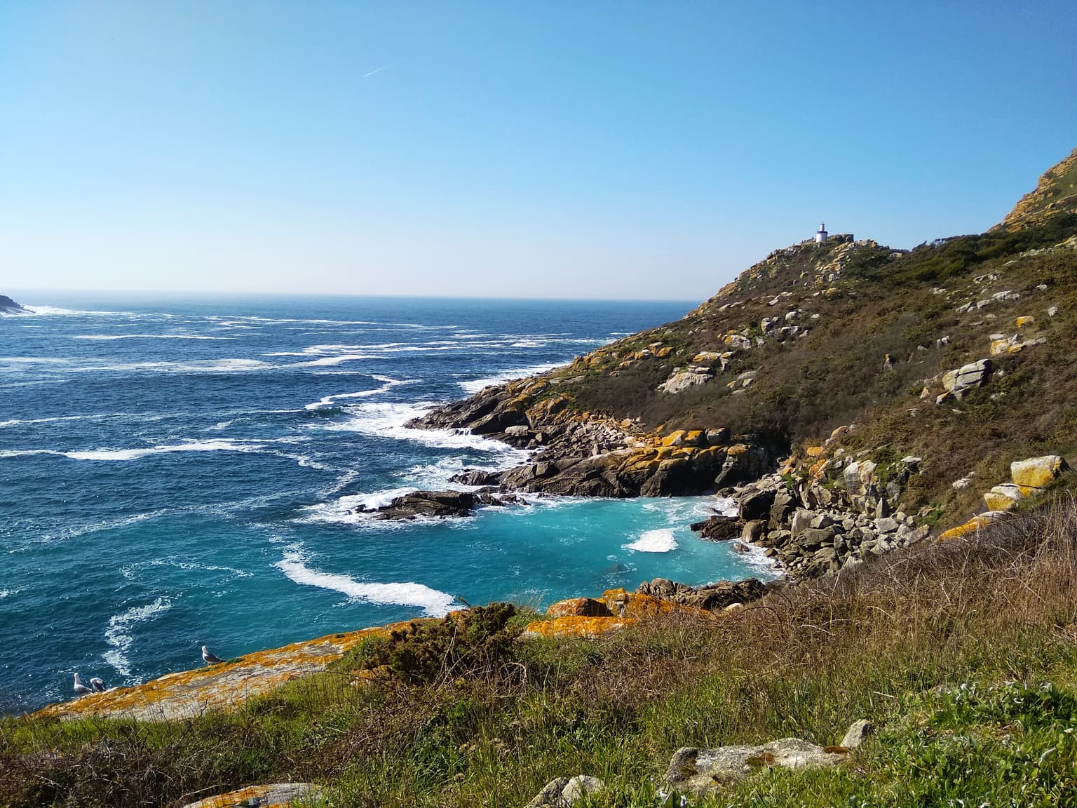 Rocky coastal landscape with turquoise ocean waters, white surf, and green vegetation on a sunny day
