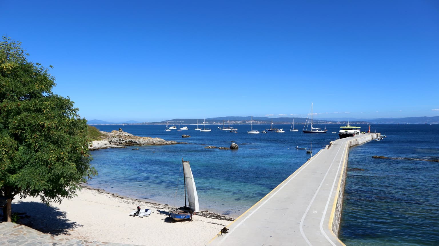Concrete pier extending into blue sea with sailboats docked, sandy beach, and tree on left under clear blue sky