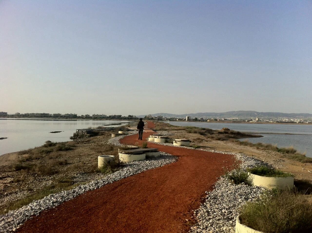 Person walking on red gravel boardwalk surrounded by water, with concrete planters and vegetation along the sides