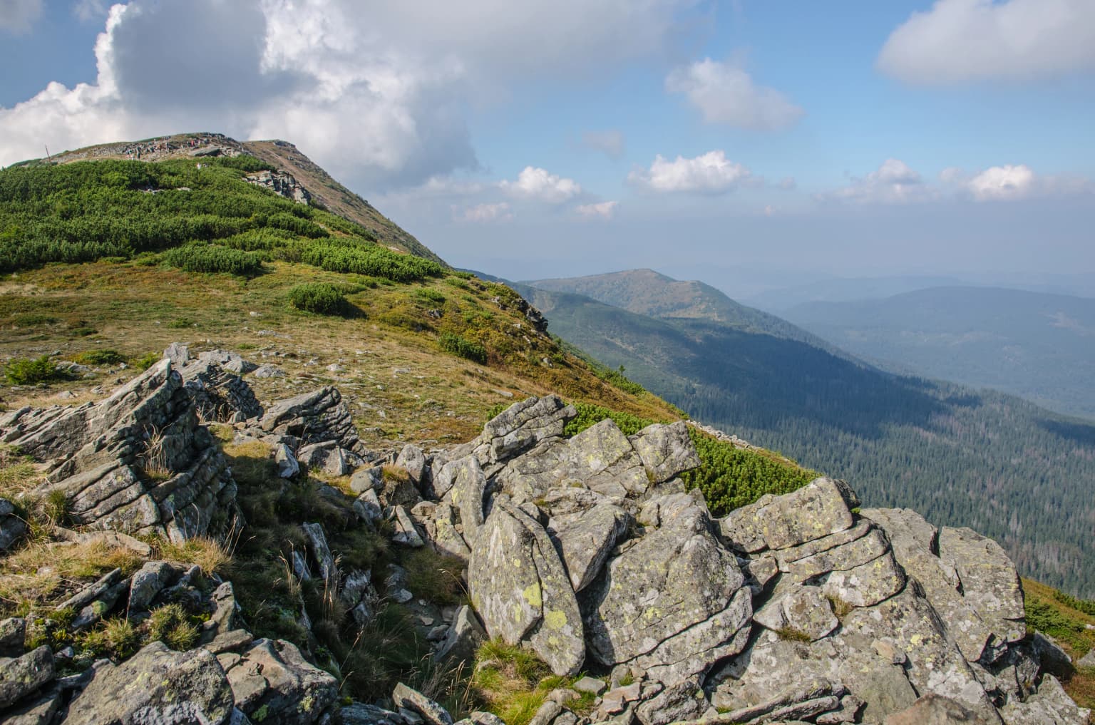 Rocky terrain with scattered boulders and patches of grass and shrubs, mountain peaks in the background under a blue sky with clouds