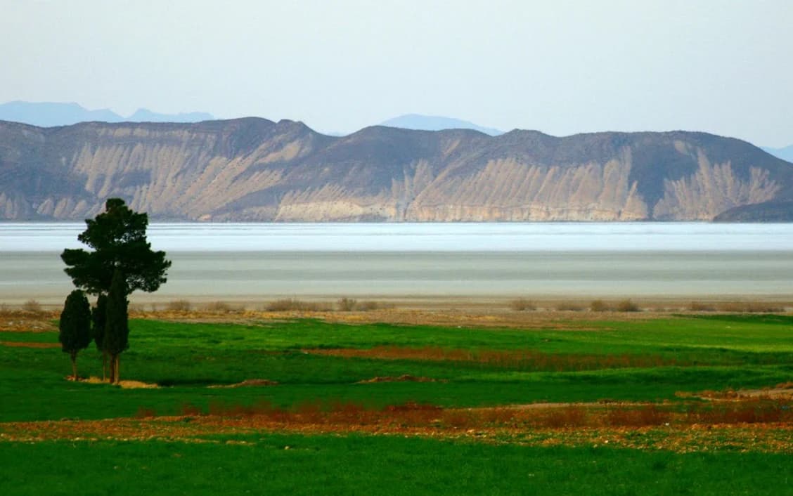 Vista del paisaje de un gran lago con campos agrícolas verdes en primer plano y montañas distantes bajo un cielo despejado