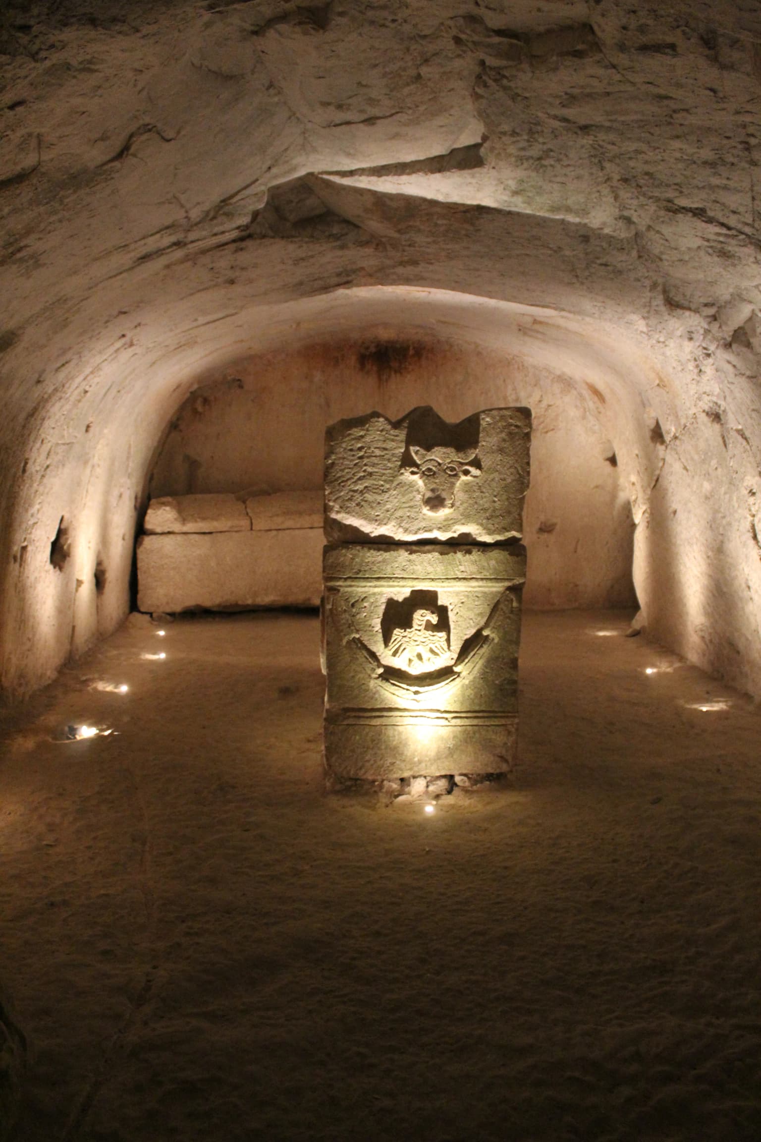 Sarcófago de piedra con decoraciones talladas, iluminado por pequeñas luces en el interior de una cámara subterránea con paredes de piedra arqueadas