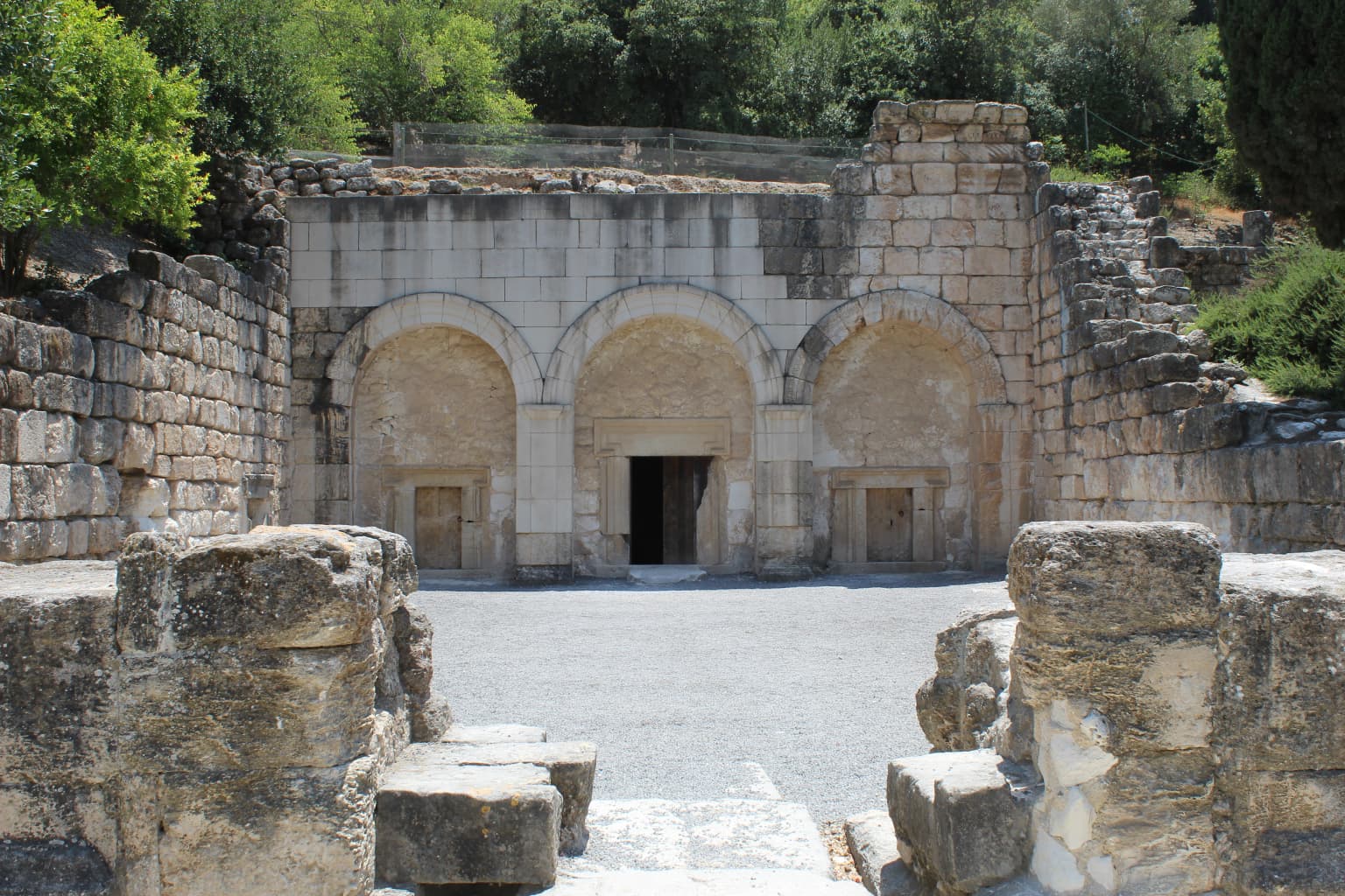 Arcos de piedra y muros reconstruidos de la entrada de una cueva funeraria rodeados de ruinas antiguas y vegetación