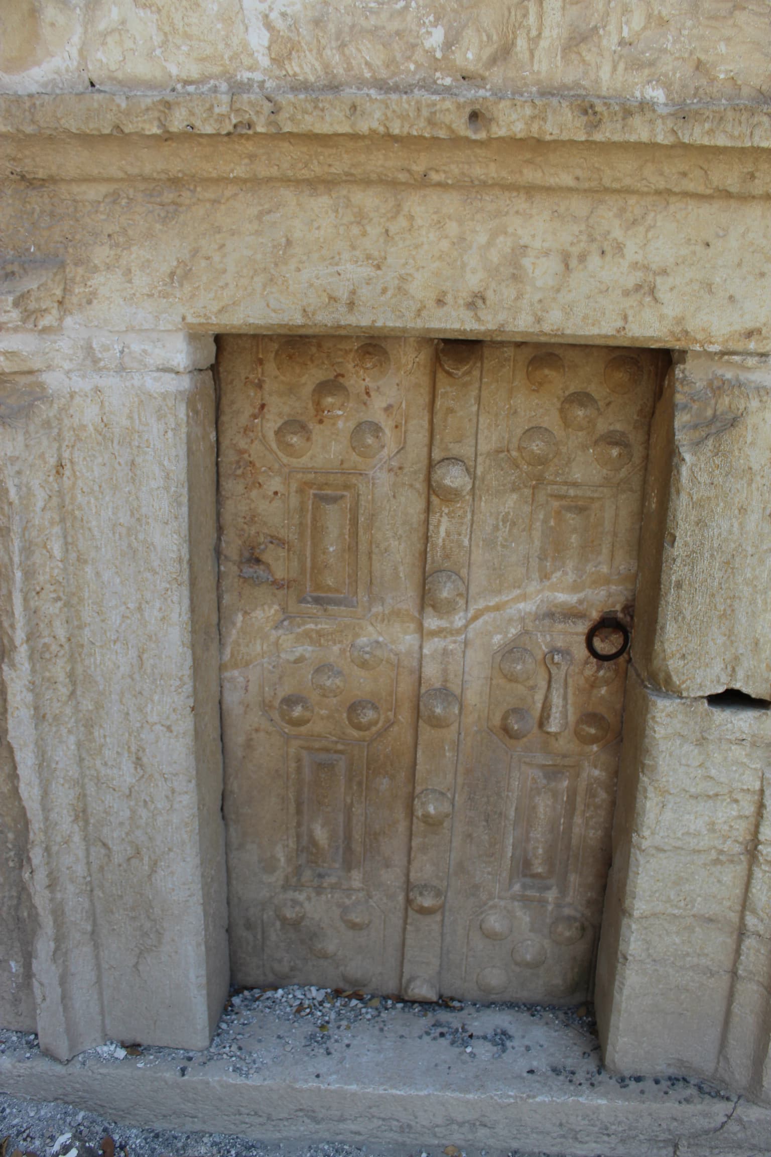 Puerta de piedra con relieves que incluyen pomos redondos y paneles rectangulares, enmarcada en piedra con un tirador anular circular.