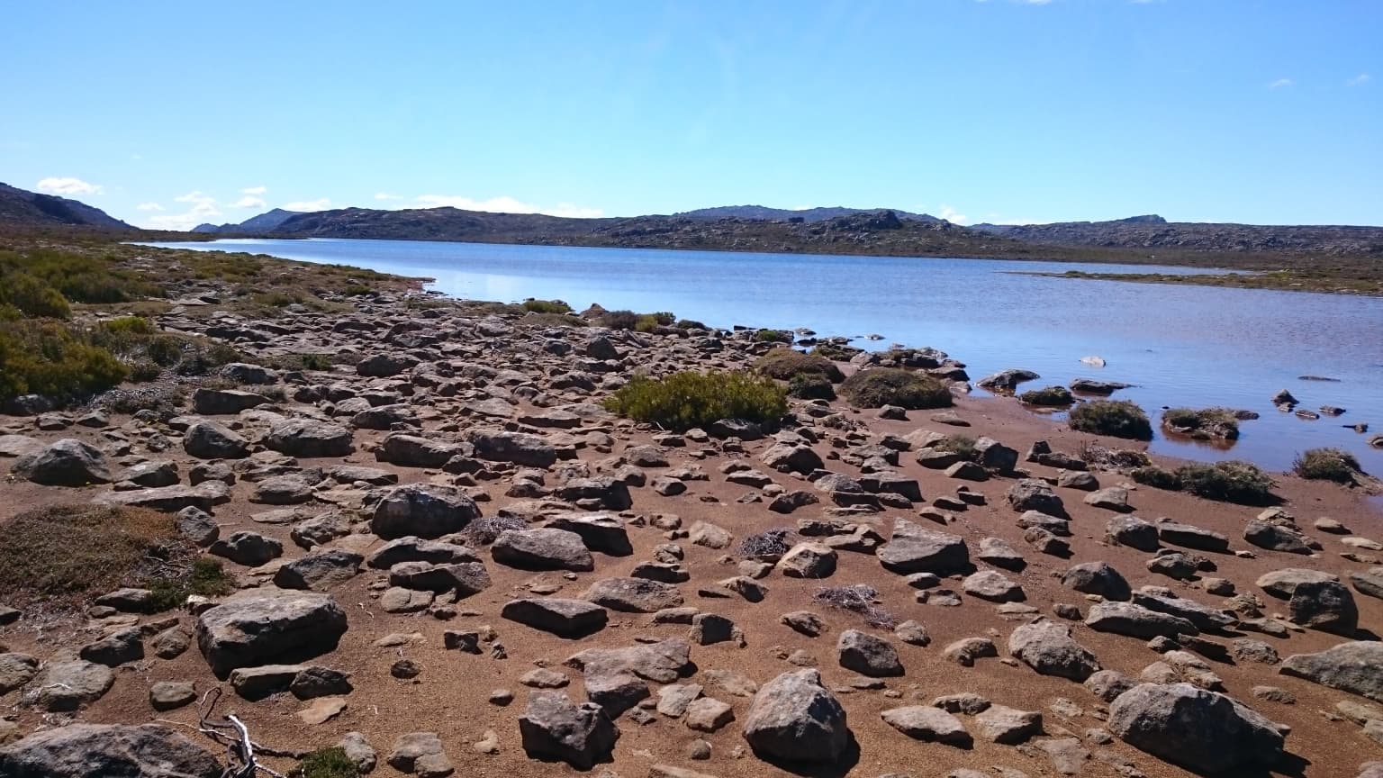Un lago rodeado de terreno rocoso con vegetación escasa bajo un cielo azul claro
