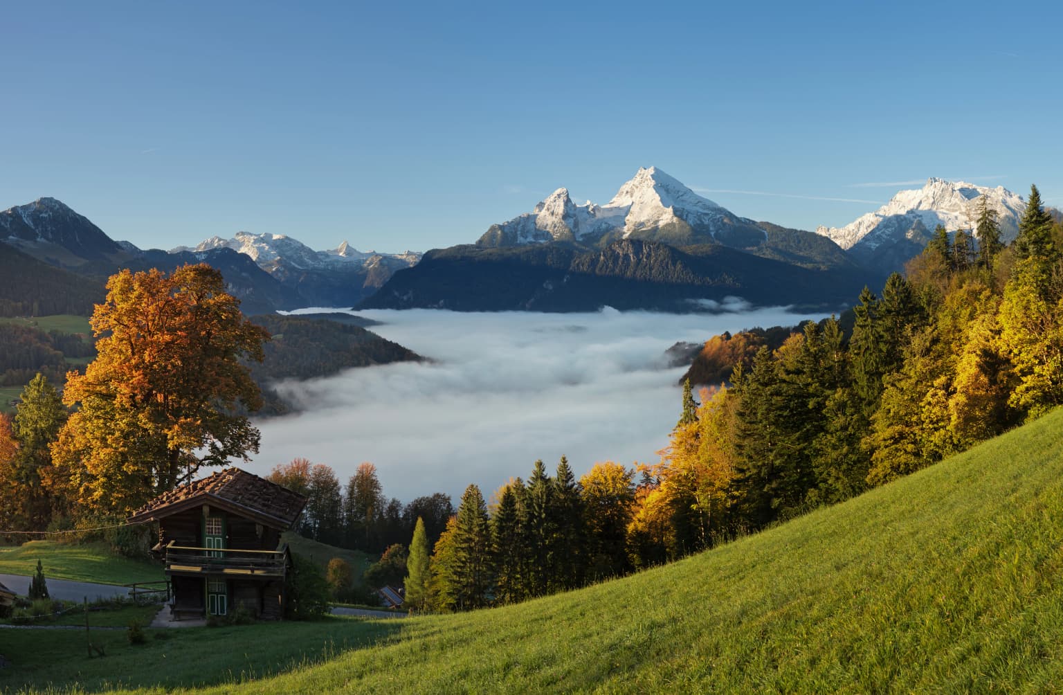 Chalet on grassy hillside with autumn trees, misty valley, and mountain peaks