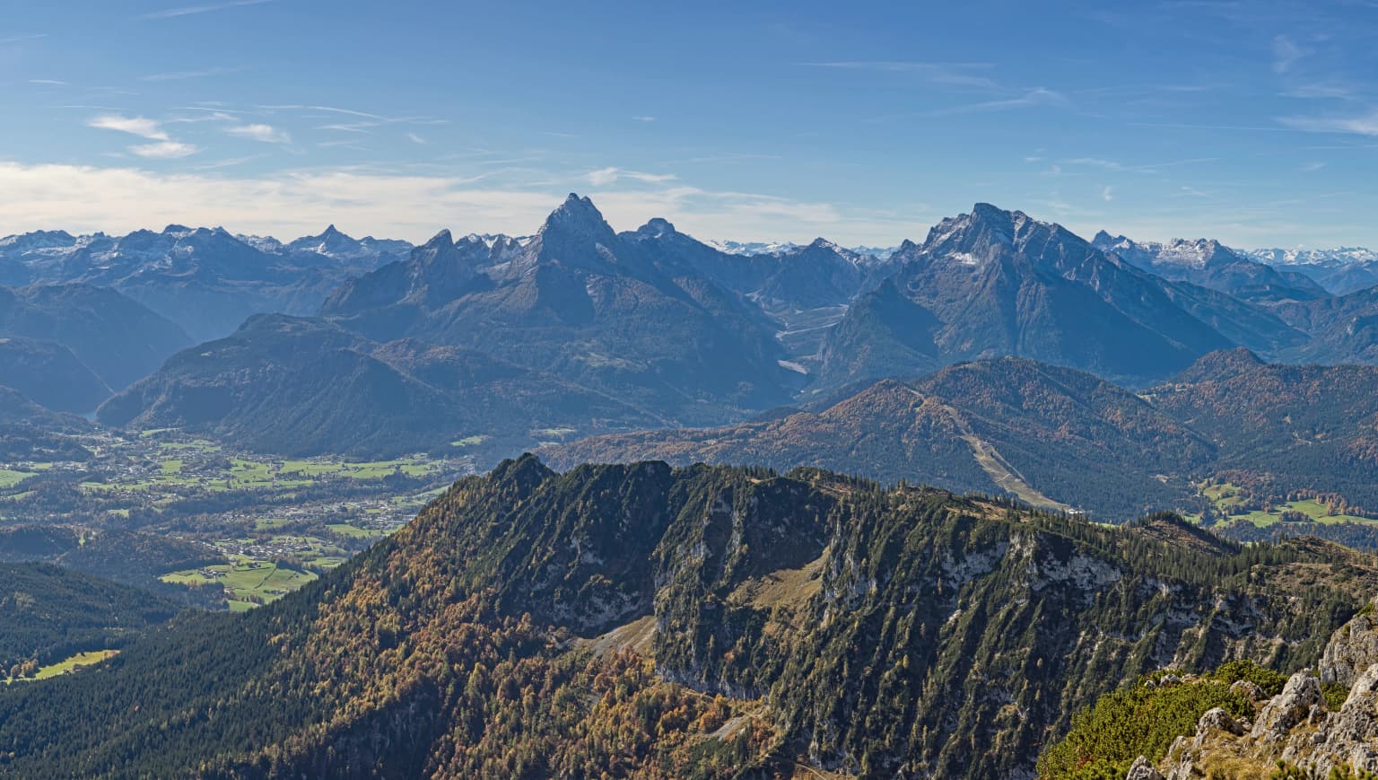Panoramic mountain landscape with multiple peaks, valleys, and forested hills under a clear blue sky