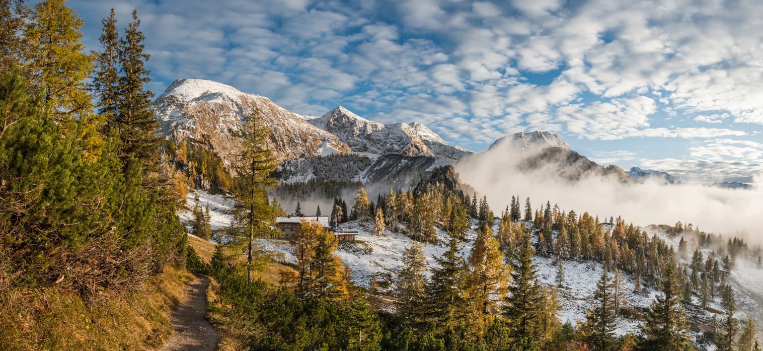 Wide landscape view of Berchtesgaden National Park with snow-covered mountain peaks, mixed forest slopes, valley mist, and clear blue sky with scattered clouds