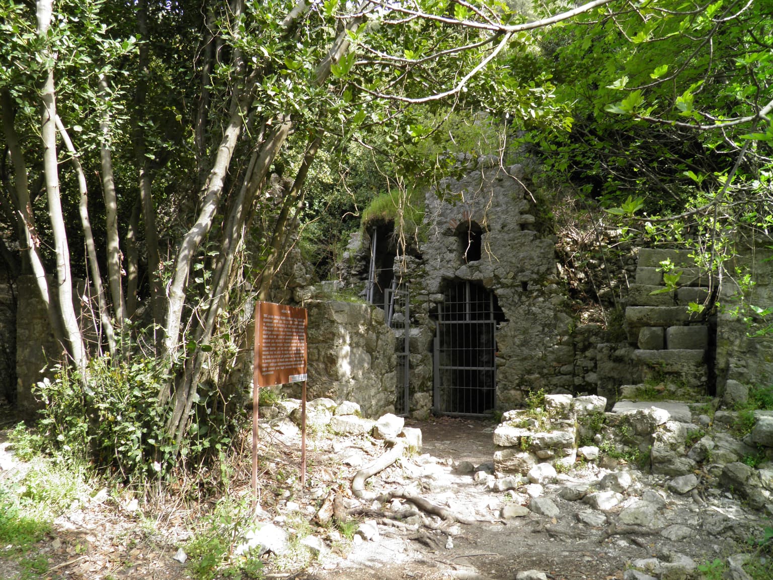 Ancient stone ruin with barred entrance surrounded by trees and vegetation, with a signboard on the left
