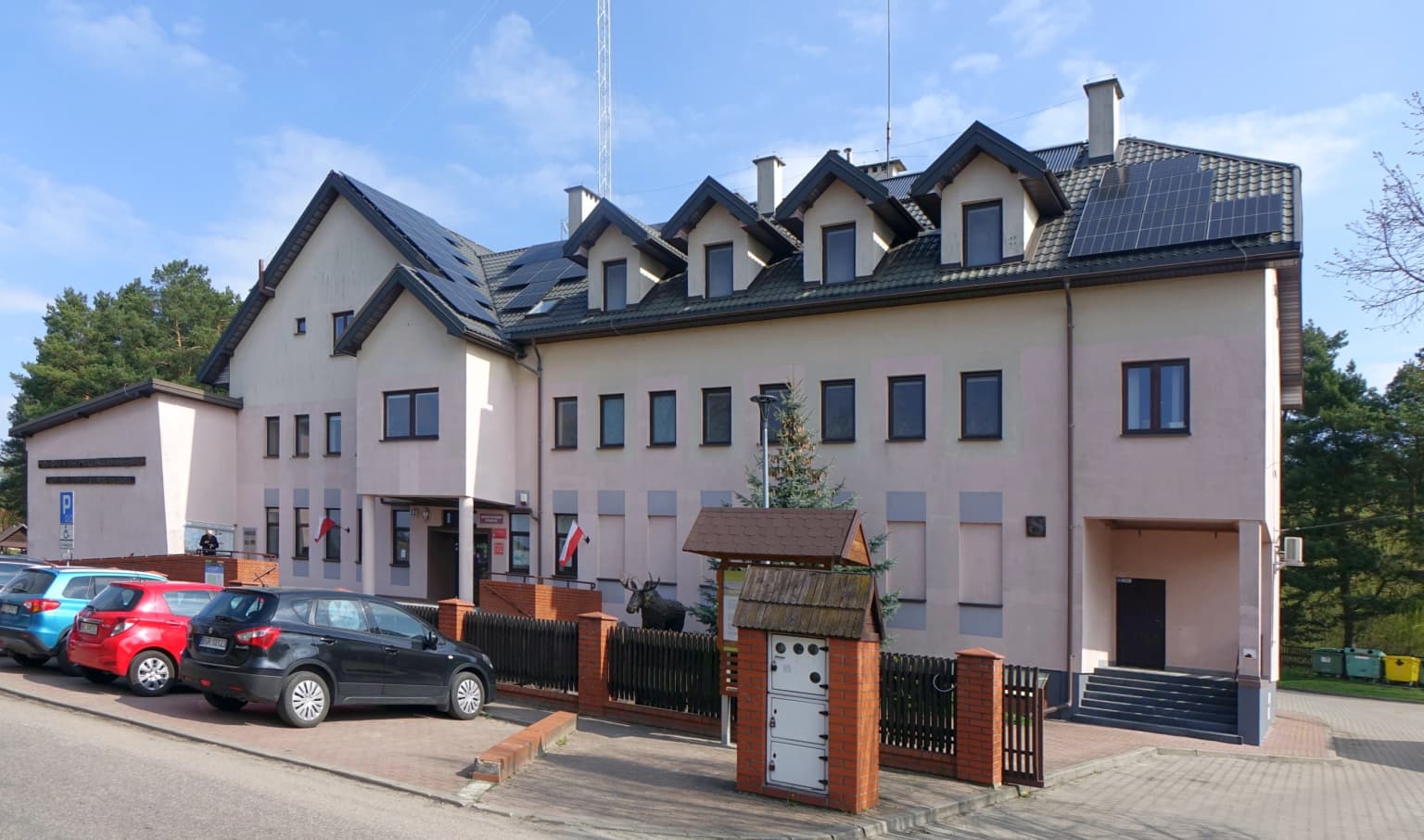 Light-colored multi-story building with dark roof, multiple windows, and a sign reading 'Biebrzański Park Narodowy' with parked cars in front