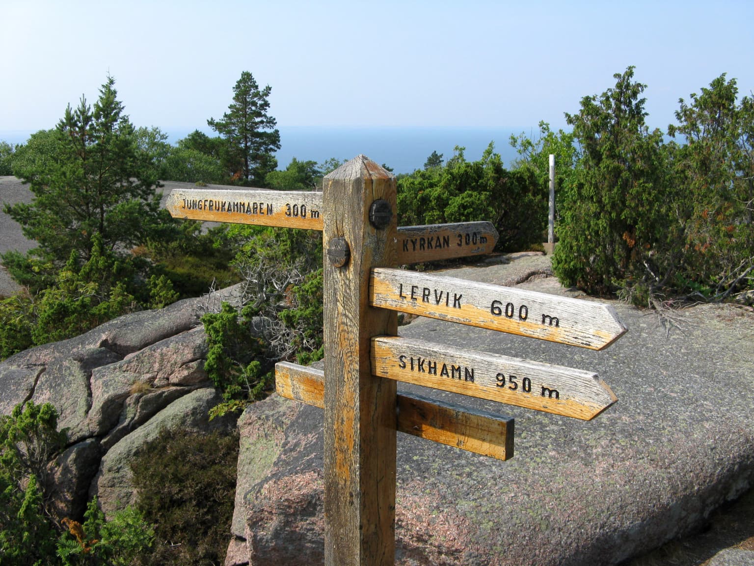 Wooden signpost with multiple directional signs displaying distances to Lervik (600 m), Sirkhamn (950 m), and other locations, set against rocky terrain with trees and distant sea view