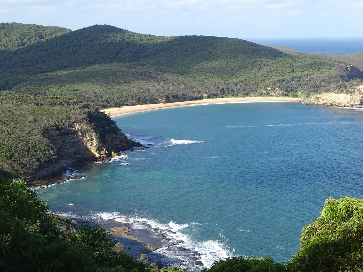 Vista al mar con playa de arena, costa rocosa, colinas boscosas y olas rompiendo contra la orilla