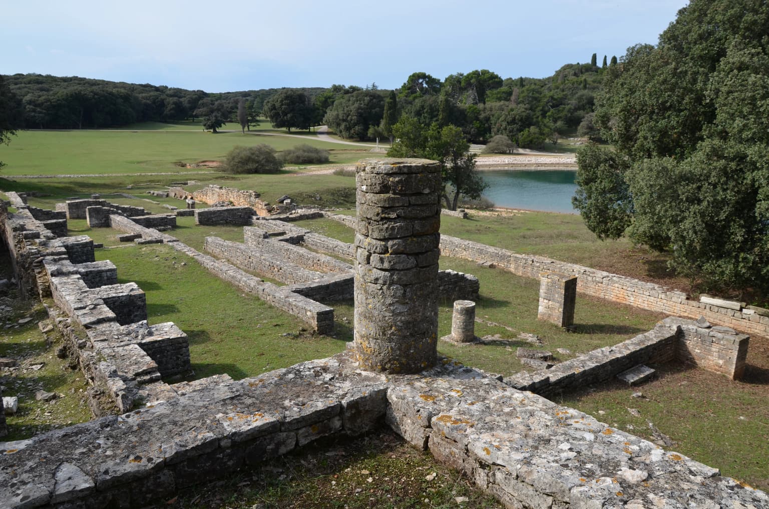 Stone ruins of ancient Roman villa structures with walls and cylindrical tower, grassy landscape, body of water, and trees in background