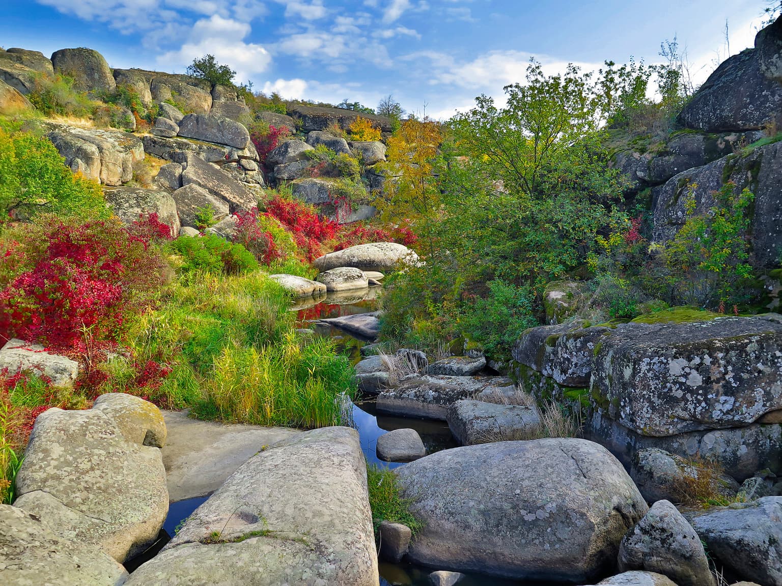Rocky canyon landscape with small stream, autumn-colored vegetation, and large boulders under partly cloudy sky