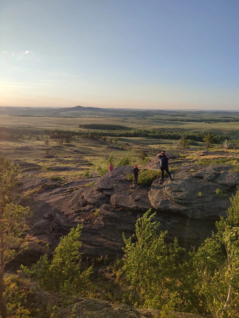 Three people standing on rocky terrain overlooking open steppe landscape with scattered trees and distant hills under clear sky
