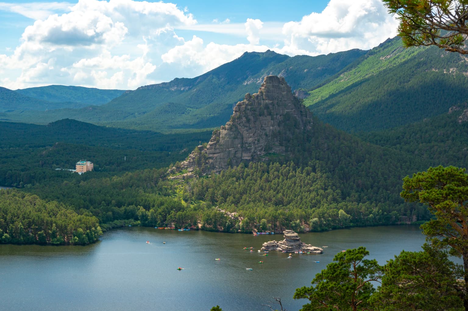 Tall rocky mountain peak with dense forest, lake, and distant mountains under a partly cloudy sky