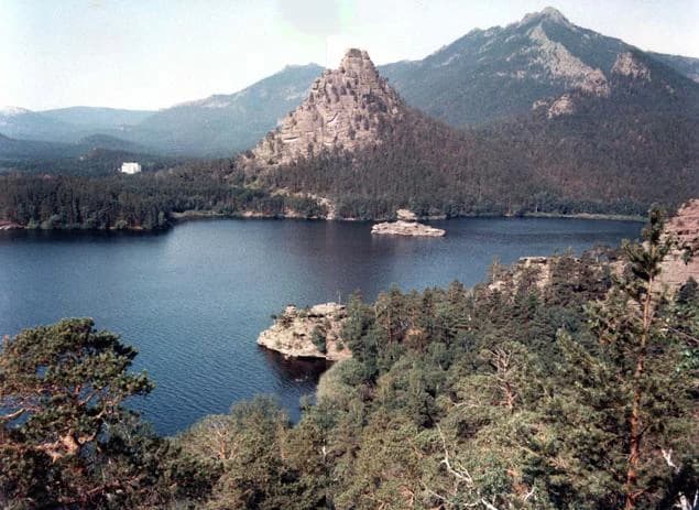 A large lake bordered by dense forest and rugged mountains with a distinctive rocky hill in the center