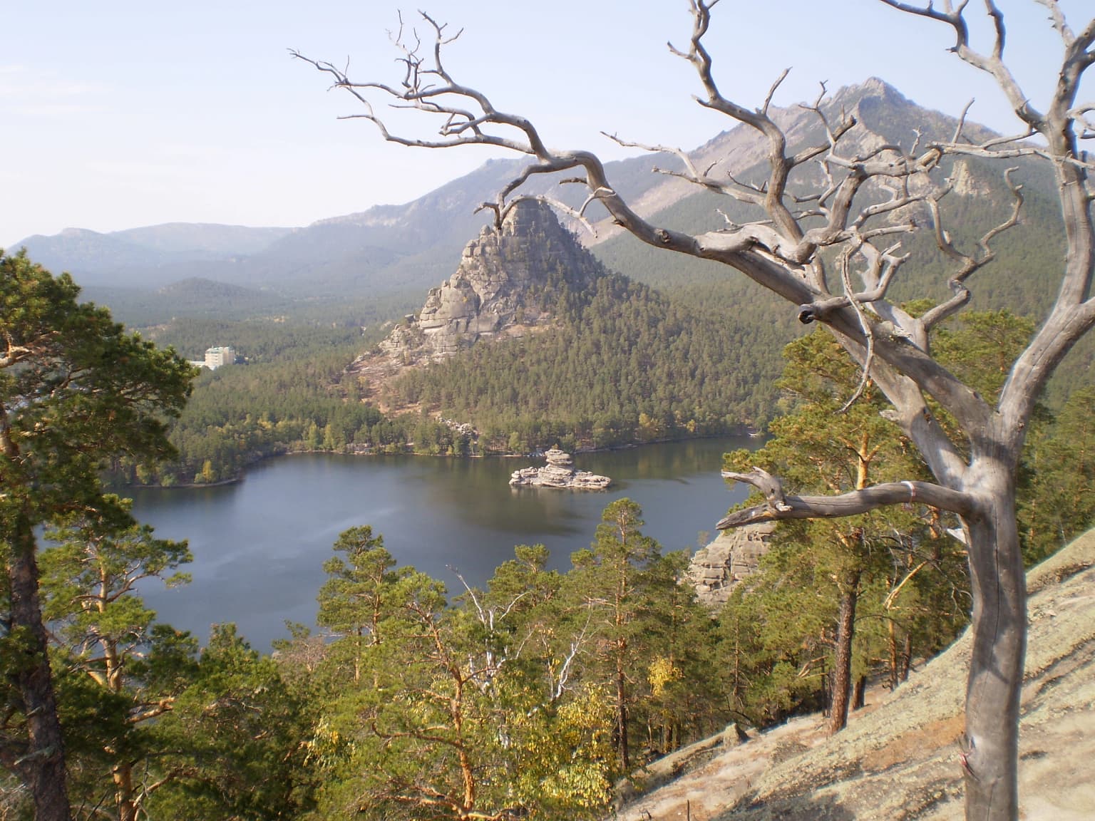 Lake Borovoye with forested mountains in the background and a distinctive rock formation, viewed from a hillside with trees in the foreground