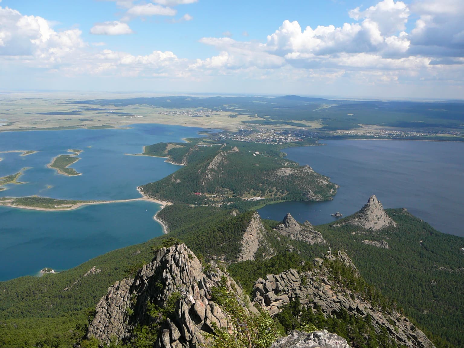 Panoramic landscape view showing two large lakes connected by a narrow isthmus, surrounded by rocky mountain peaks and dense forest under a partly cloudy sky