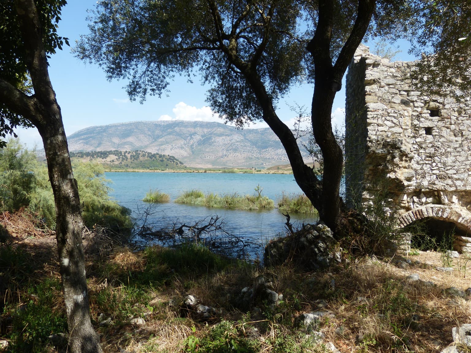 Stone ruin structure by a lake with trees, vegetation, and mountains in the background
