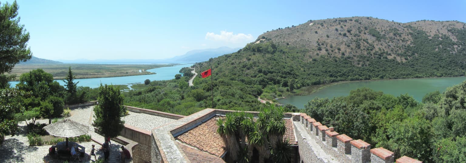 Panoramic view of Butrint National Park with stone ruins, trees, a lake, hills, and a red flag