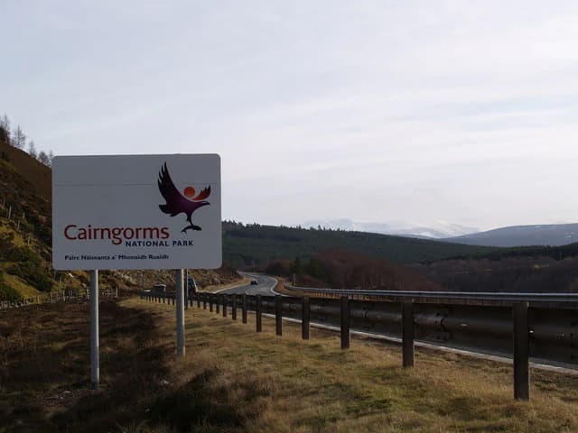White Cairngorms National Park sign with bird logo, A9 road with guardrails, grassy hillside, and mountain range in background