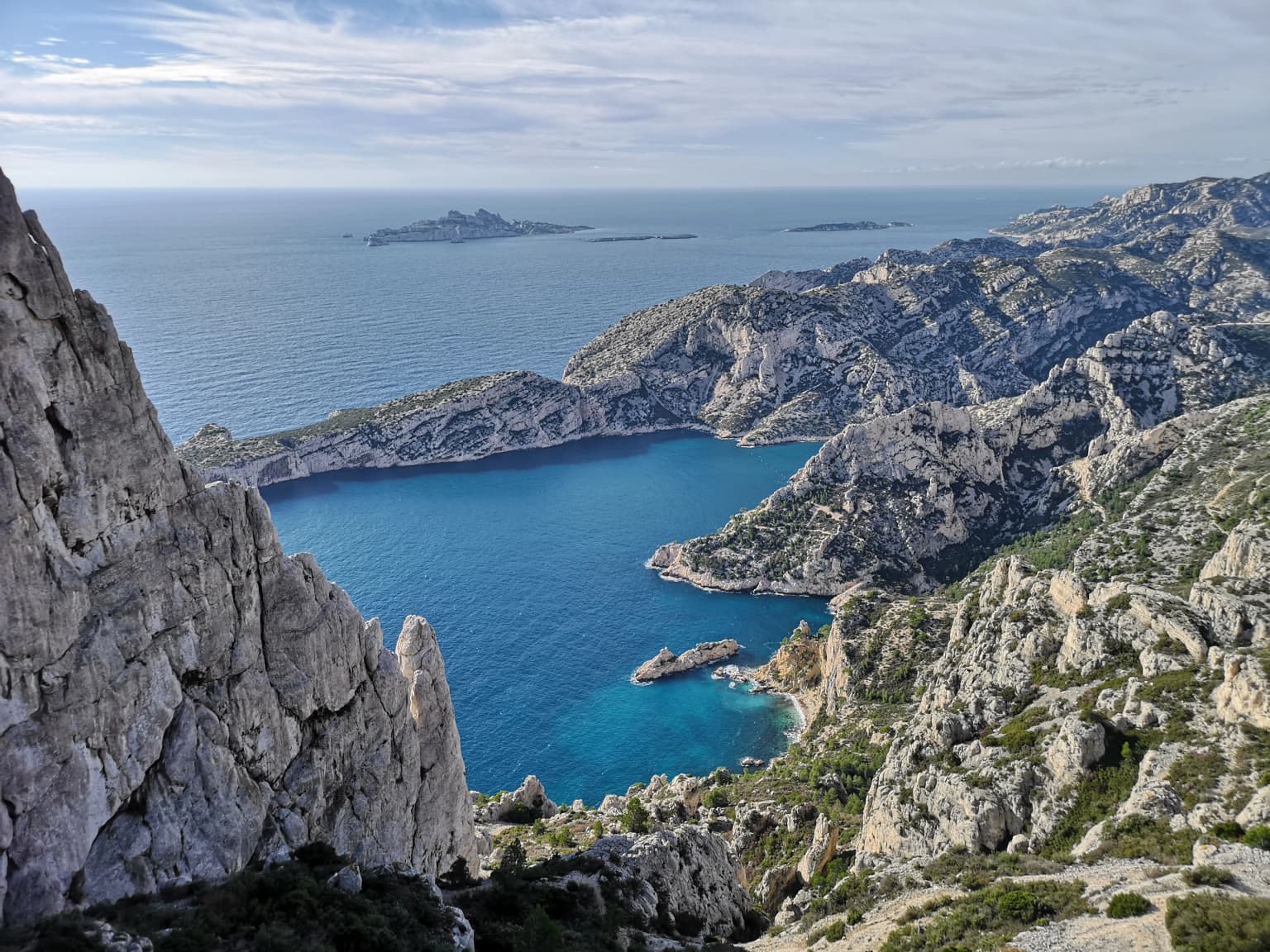 Rocky coastal cliffs overlooking turquoise water coves, distant islands, and mountainous terrain under a partly cloudy sky