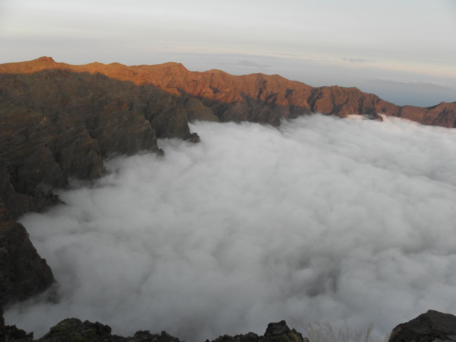 Panoramic view of a volcanic caldera with clouds filling the basin and rocky mountain peaks surrounding it under a partly cloudy sky