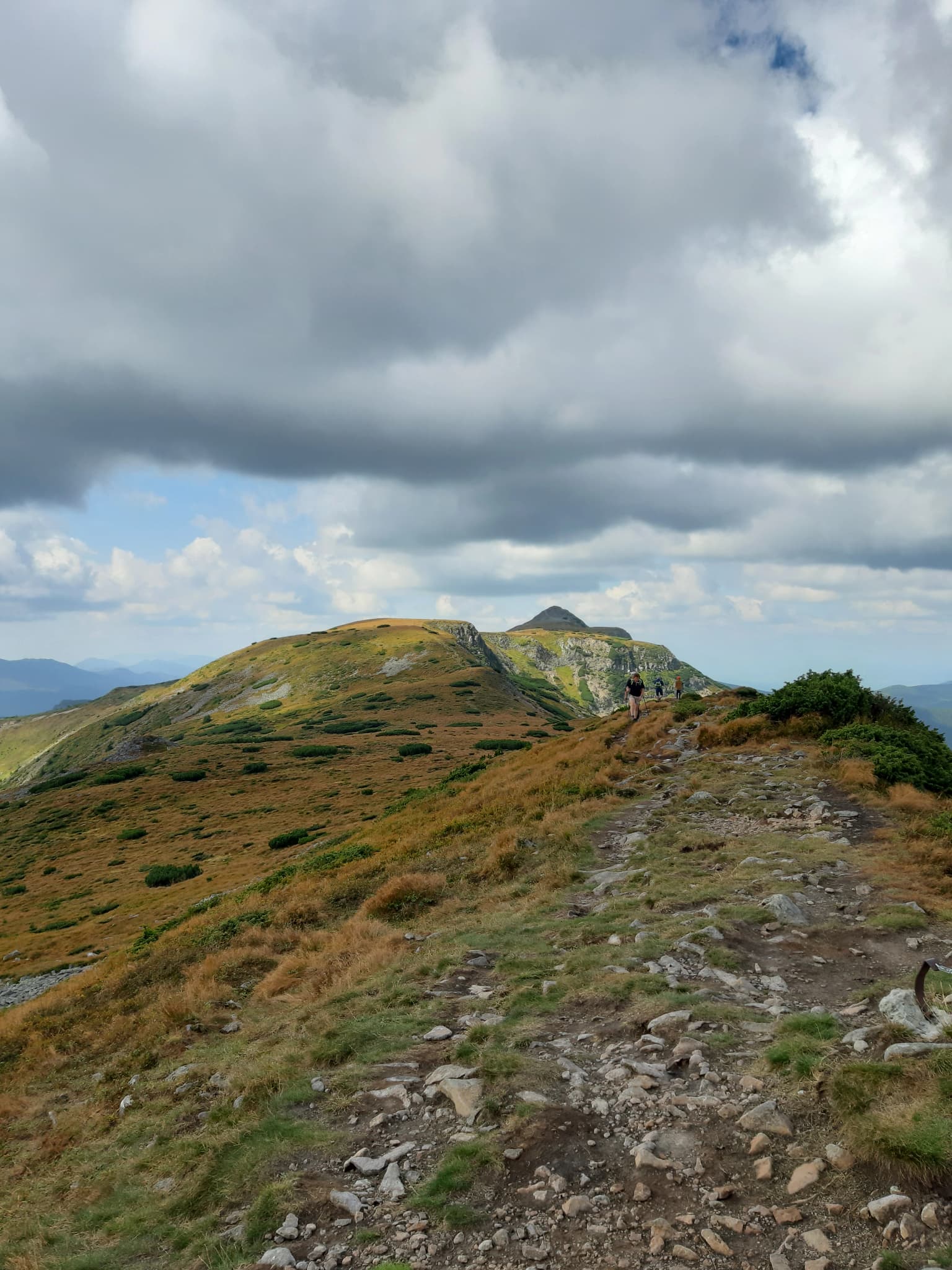 Rocky mountain trail ascending through grassy slopes with a hiker in the distance under a partly cloudy sky